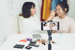 © sutthinon602 - Two young girls doing video recording by smartphone camera. Asian woman make up artist applying powder on female blogger face with cosmetic products table in living room. Beauty ladies live stream