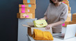 © sutthinon602 - Young business woman working selling online. Entrepreneur owner using Laptop or tablet taking receive and checking online purchase shopping order to preparing pack product box. Online shopping