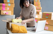 © sutthinon602 - Young business woman working selling online. Entrepreneur owner using Laptop or tablet taking receive and checking online purchase shopping order to preparing pack product box. Online shopping