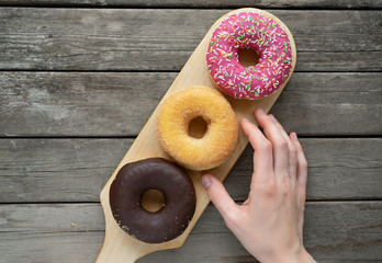 Glazed three donuts in a row on a wooden board background with blank space. Woman's hand reaches for a yellow handwriting, wants to taste