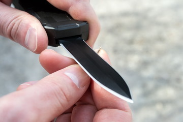 Closeup view of male hands sharpening a small thin wooden stick with a black pocket out the front knife with a sharp blade. Blurred background