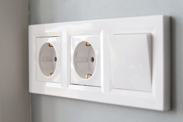 A closeup diagonal view of a group of white european electrical outlets and a switch located on a painted gray wall