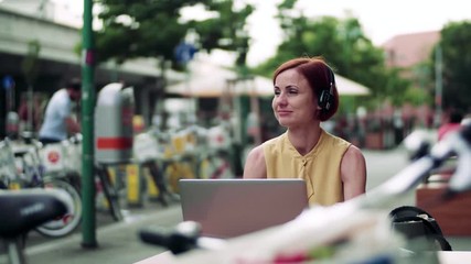 Wall Mural - Young businesswoman with headphones and laptop sitting in cafe outdoors in city.