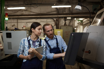 Bearded young engineer working on machine and showing something to young woman with tablet pc in the plant