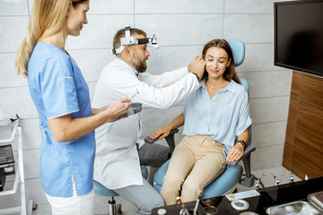 Wall Mural - Senior otolaryngologist examining ear of a young patient with female assistant in ENT office, nurse giving medical tools