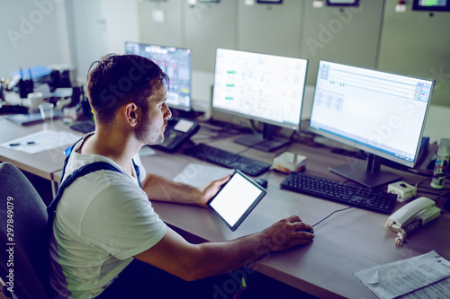 Fotografía Hardworking plant worker sitting in control room, holding tablet and using computer for monitoring