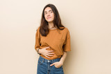Fototapeta Panele - Young brunette woman against a beige background touches tummy, smiles gently, eating and satisfaction concept.