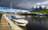 A closeup of a yacht moored at Newcastle's Quayside on the river Tyne with the Baltic Art Museum, Gateshead Sage and Millennium Bridge in the distance.