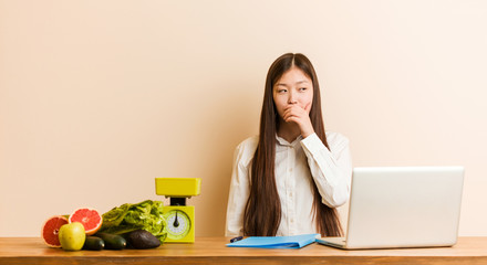 Wall Mural - Young nutritionist chinese woman working with her laptop thoughtful looking to a copy space covering mouth with hand.