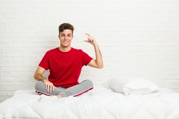 Wall Mural - Young teenager student man on the bed holding something little with forefingers, smiling and confident.