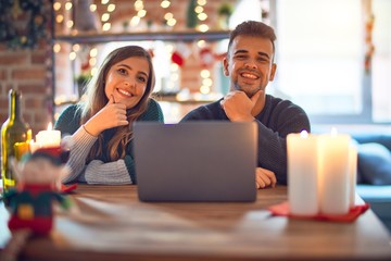 Sticker - Young beautiful couple sitting using laptop around christmas decoration at home looking confident at the camera smiling with crossed arms and hand raised on chin. Thinking positive.