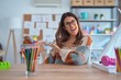 © Krakenimages.com - Young beautiful teacher woman wearing sweater and glasses sitting on desk at kindergarten Inviting to enter smiling natural with open hand