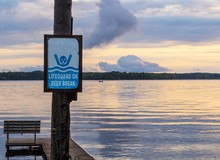A Humorous Sign On The Beach Announces That The Lifeguard Is On A Beer Break, With A Lake At Sunset In The Background