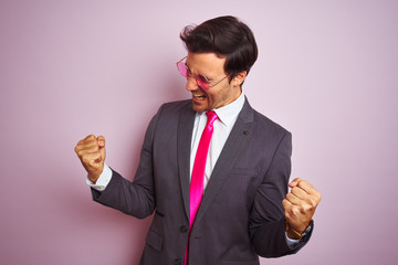 Wall Mural - Young handsome businessman wearing suit and sunglasses over isolated pink background very happy and excited doing winner gesture with arms raised, smiling and screaming for success. 