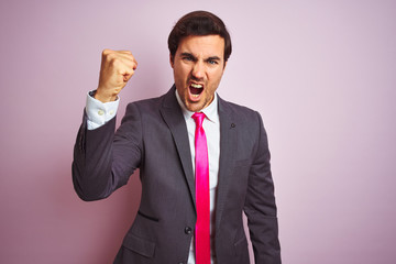 Wall Mural - Young handsome businessman wearing suit and tie standing over isolated pink background angry and mad raising fist frustrated and furious while shouting with anger. Rage and aggressive concept.