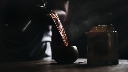 dark vintage background of blurred man pouring hot yellow chinese tea to teacup with old traditional chinese tea pot on table