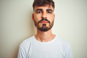 Sticker - Young man with tattoo wearing t-shirt standing over isolated white background with serious expression on face. Simple and natural looking at the camera.