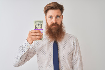 Young redhead irish businessman holding dollars standing over isolated white background with a confident expression on smart face thinking serious