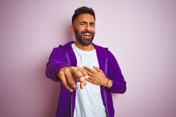 Wall Mural - Young indian man wearing purple sweatshirt standing over isolated pink background Laughing of you, pointing finger to the camera with hand over mouth, shame expression