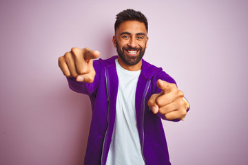 Young indian man wearing purple sweatshirt standing over isolated pink background pointing to you and the camera with fingers, smiling positive and cheerful