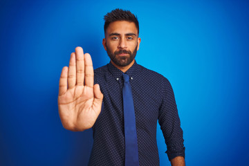 Poster - Young indian businessman wearing elegant shirt and tie standing over isolated blue background doing stop sing with palm of the hand. Warning expression with negative and serious gesture on the face.