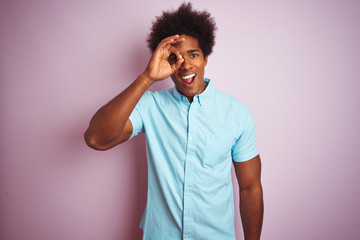 Poster - Young american man with afro hair wearing blue shirt standing over isolated pink background doing ok gesture with hand smiling, eye looking through fingers with happy face.