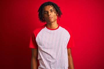 Wall Mural - Afro man with dreadlocks wearing striped t-shirt standing over isolated red background with serious expression on face. Simple and natural looking at the camera.