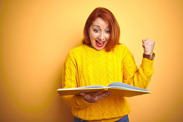 Young redhead student woman reading book standing over yellow isolated background screaming proud and celebrating victory and success very excited, cheering emotion