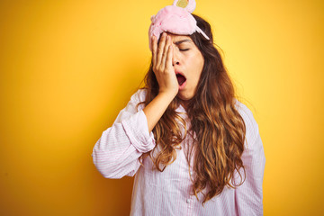 Wall Mural - Young woman wearing pajama and sleep mask standing over yellow isolated background Yawning tired covering half face, eye and mouth with hand. Face hurts in pain.