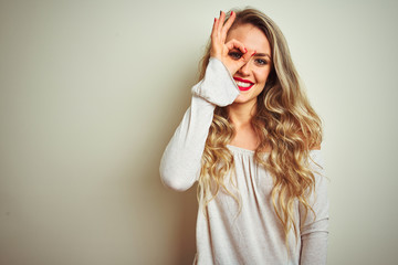 Canvas Print - Young beautiful woman standing over white isolated background doing ok gesture with hand smiling, eye looking through fingers with happy face.