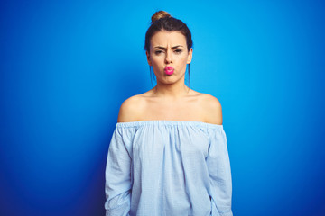 Sticker - Young beautiful woman wearing bun hairstyle over blue isolated background puffing cheeks with funny face. Mouth inflated with air, crazy expression.