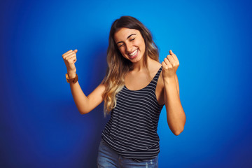 Poster - Young beautiful woman wearing stripes t-shirt over blue isolated background very happy and excited doing winner gesture with arms raised, smiling and screaming for success. Celebration concept.