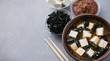 Bowl with miso soup, wakame seaweed, miso pasta, tofu and chopsticks on a gray background.
