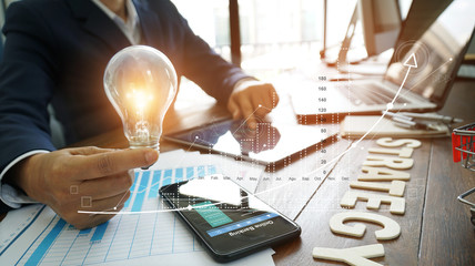 Poster - Businessman hand holding lightbulb with business documents and strategy word and banking data on smart phone and digital tablet with graph business diagram, finance ideas and strategy concept