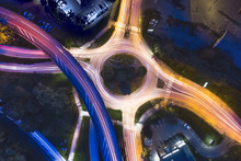 Transportation At Night With Commuting Vehicle Traffic Creating Light Trails On The Road With Motion Blur, Roundabout And Motorway Interchange In City, Drone Aerial Photography With Long Exposure