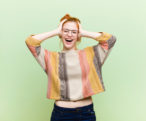 young pretty red head woman raising hands to head, open-mouthed, feeling extremely lucky, surprised, excited and happy against green wall