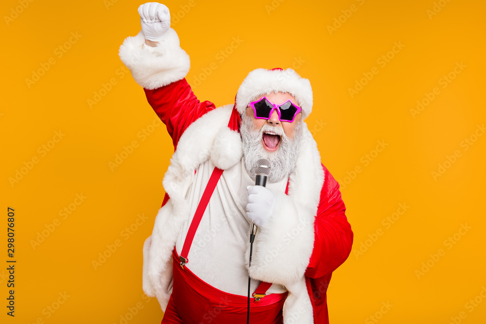 Grey haired stylish christmas grandfather in red hat cap celebrate x-mas party hold microphone sing noel carols feel funky with big belly wear suspenders isolated over yellow color background - obrazy, fototapety, plakaty 