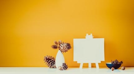 Composition of dried flowers, thorns in light vase, pine cones and blank canvas on a stand on ocher color wall background. Simple, natural home interior decor. Minimalism. Wide banner. Copy space.