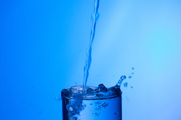 Wall Mural - Chorro de agua llenando y rebosando un vaso de agua de cristal o plástico transparente sobre fondo azul