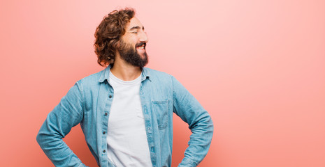Wall Mural - young bearded crazy man looking happy, cheerful and confident, smiling proudly and looking to side with both hands on hips against flat color wall
