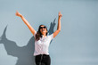 © Alexandra Coelho/ADDICTIVE STOCK - Carefree beautiful woman in casual outfit and headphones on neck standing with hands up beside blue wall of building on city street