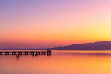 Silhouettes Of Distant People Sitting On Pier Near Tranquil Lake Against Cloudless Sunset Sky