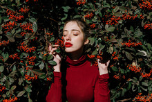 Young Female With Closed Eyes Standing Amidst Green Shrub Branches With Red Berries On Sunny Day In Garden