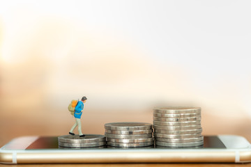 Travel Planning, Money and technology concept. Close up of traveler miniature figure with backpack walking on top stack of coins on smart mobile phone with copy space.