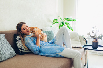 Playing with cat at home. Young woman sitting on couch teasing and hugging pet.