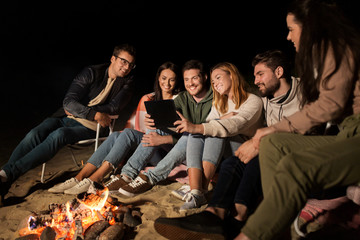 Sticker - leisure and people concept - group of smiling friends sitting at camp fire with tablet pc computer on beach at night