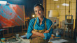 © Gorodenkoff - Portrait of Talented Young Female Artist Dirty with Paint, Wearing Apron, Crosses Arms while Holding Brushes, Looks at Camera with Smile. Authentic Creative Studio with Large Canvas Tools Everywhere