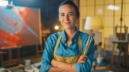 Fototapete - Talented Young Female Artist Dirty with Paint, Wearing Apron, Crosses Arms while Holding Brushes, Looks at the Camera with a Smile. Authentic Creative Studio with Large Canvas and Tools Everywhere 