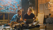 © Gorodenkoff - Couple of Artists Sitting in their Studio Use Laptop Computer, Working on a Project, Talking and Smiling. Painter and Her Partner Doing Research. Authentic Workshop with Oil Paintings and Tools