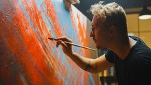 Portrait Of Artist Working On Abstract Painting, Uses Paint Brush To Create Daringly Emotional Modern Picture. Dark Creative Studio Large Canvas Stands On Easel Illuminated. Side View Close-up Shot 